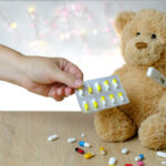 A teddy bear sits on a table with a thermometer under one arm, surrounded by loose pills and one blister pack of pills.