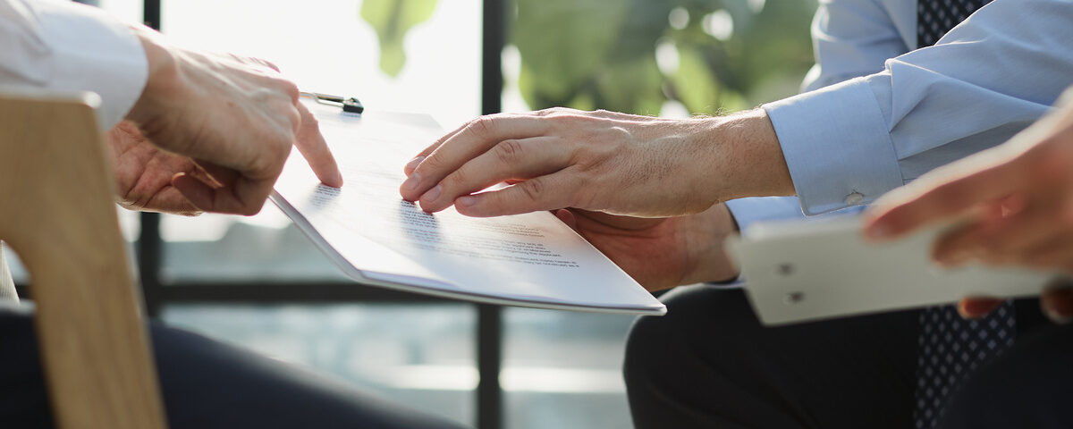 Three people’s hands are visible as they review documents on clipboards. One points to a paper on another’s clipboard.