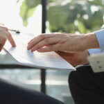 Three people’s hands are visible as they review documents on clipboards. One points to a paper on another’s clipboard.