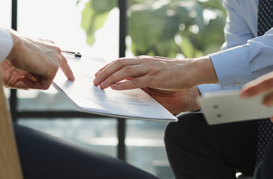 Three people’s hands are visible as they review documents on clipboards. One points to a paper on another’s clipboard.