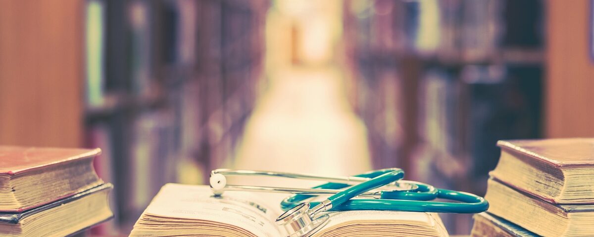 At the end of a library’s aisle, several books sit on a table. One book is open, with a stethoscope laid on top.