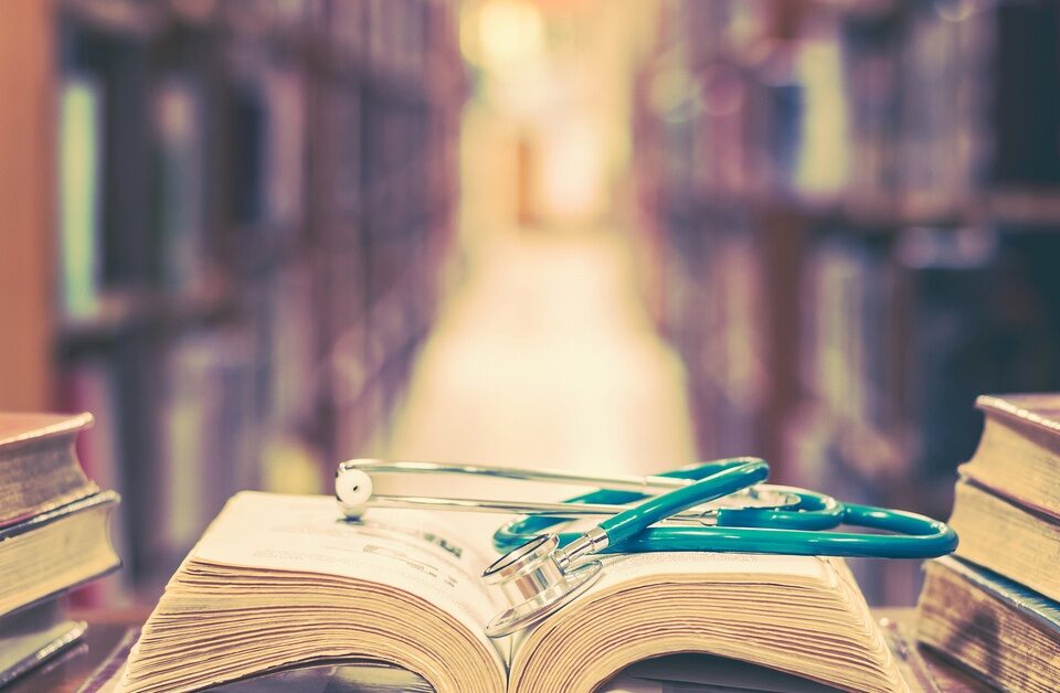 At the end of a library’s aisle, several books sit on a table. One book is open, with a stethoscope laid on top.