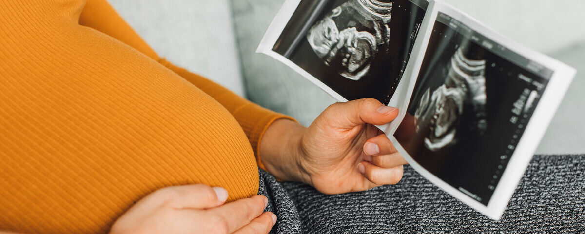 A visibly pregnant person in an orange shirt sitting with one hand on her belly while holding a sonogram in the other hand.