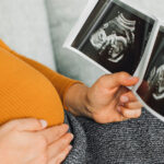 A visibly pregnant person in an orange shirt sitting with one hand on her belly while holding a sonogram in the other hand.