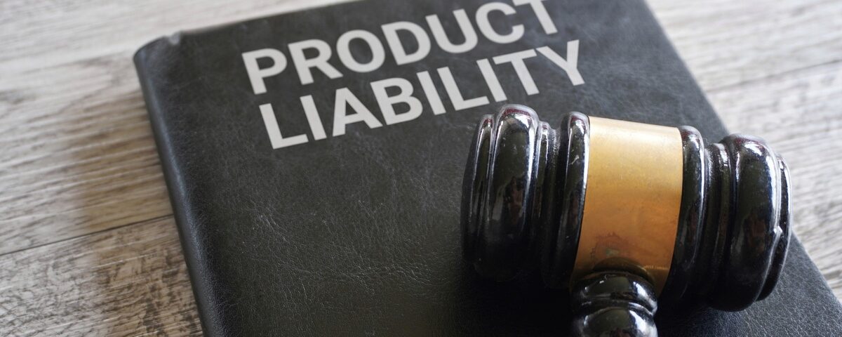 A wooden gavel lays on top of a black notebook that says “Product Liability.” The items sit on a wooden table.
