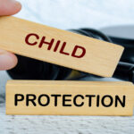 A person’s hand arranges two wooden blocks on a table with a gavel. One wooden block has the word “Child” on it and the other has the word “Protection” on it.
