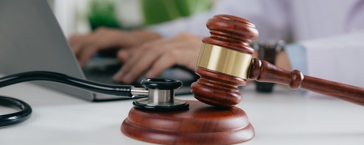 A wooden gavel and a stethoscope sit on a table in the foreground. In the background, a person works at a laptop.