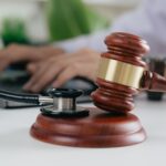 A wooden gavel and a stethoscope sit on a table in the foreground. In the background, a person works at a laptop.