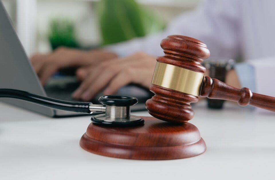 A wooden gavel and a stethoscope sit on a table in the foreground. In the background, a person works at a laptop.