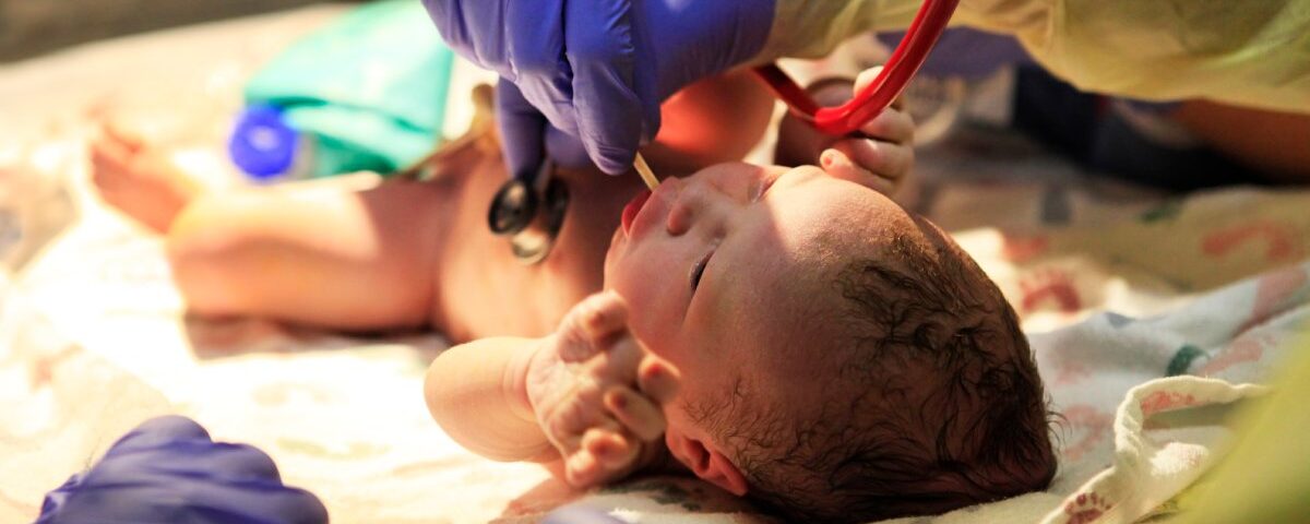 A newborn baby lying on their back beneath a light. People wearing surgical gloves check the baby's vital signs.