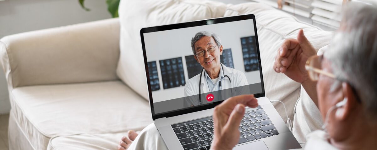 A man in a white shirt sits on a couch with a laptop in his lap. He is on a video call with his doctor.