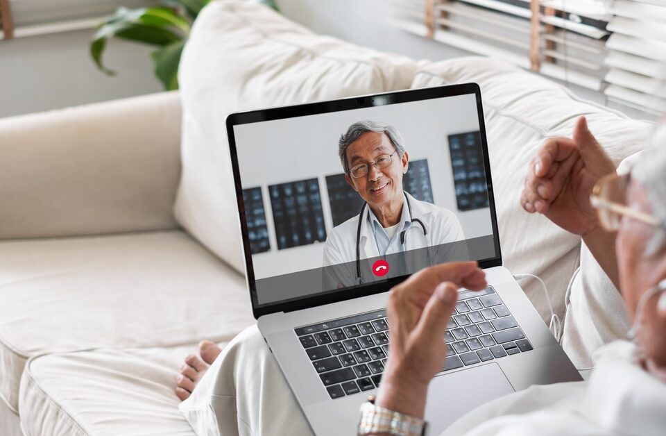 A man in a white shirt sits on a couch with a laptop in his lap. He is on a video call with his doctor.