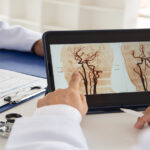 A person at a table gestures at a digital tablet that displays images from a brain scan. Another person fills out paperwork.