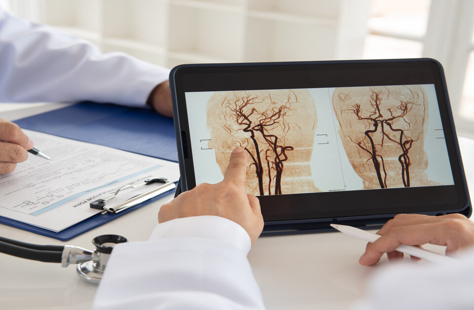 A person at a table gestures at a digital tablet that displays images from a brain scan. Another person fills out paperwork.