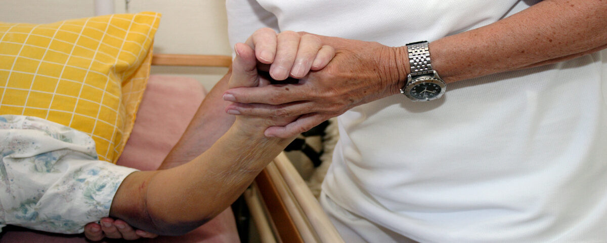 A person in white scrubs uses their hands to support the arm of a patient who is lying in a hospital bed.