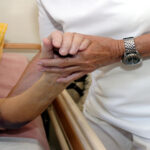 A person in white scrubs uses their hands to support the arm of a patient who is lying in a hospital bed.