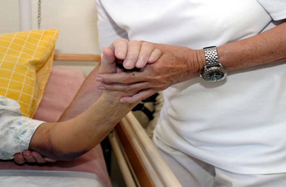 A person in white scrubs uses their hands to support the arm of a patient who is lying in a hospital bed.