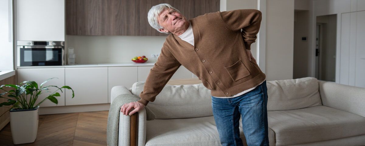 A grey-haired man in a brown cardigan stands near his couch. He holds his lower back and winces in pain.