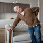 A grey-haired man in a brown cardigan stands near his couch. He holds his lower back and winces in pain.