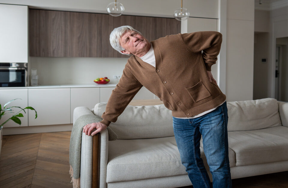 A grey-haired man in a brown cardigan stands near his couch. He holds his lower back and winces in pain.