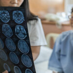 A doctor holds up a sheet with a series of MRI images of the brain and shows it to the patient, a woman with glasses.