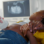 A woman lies on an exam table, getting an ultrasound of her unborn child. A man holds her hand as they watch the screen.