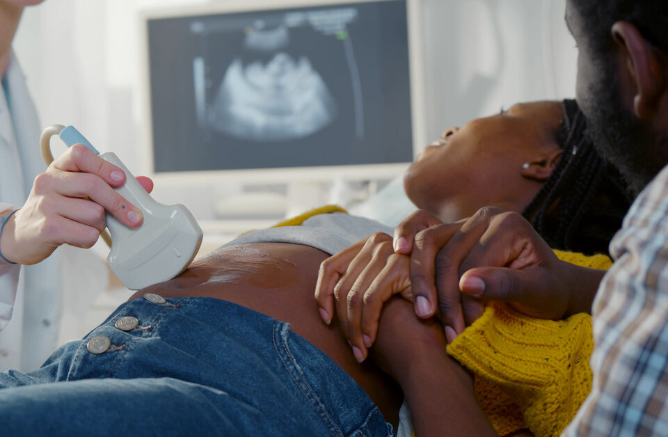 A woman lies on an exam table, getting an ultrasound of her unborn child. A man holds her hand as they watch the screen.