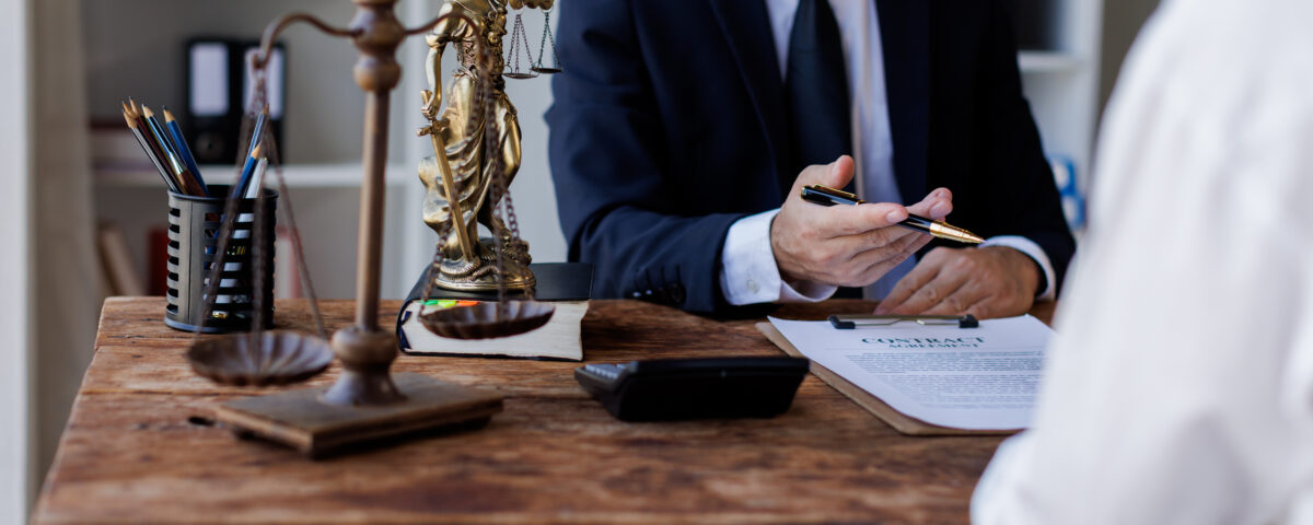 A legal expert in a black suit sits at their desk in front of a client. A scale figure and gavel sit on the desk corner.