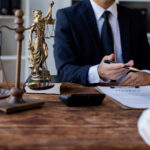 A legal expert in a black suit sits at their desk in front of a client. A scale figure and gavel sit on the desk corner.