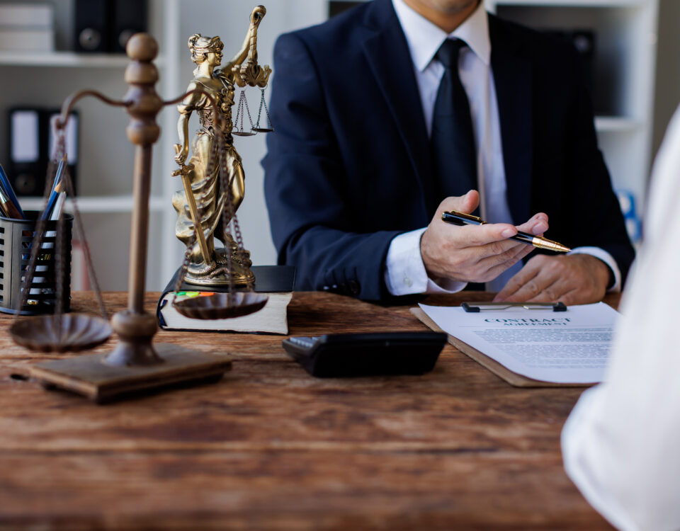 A legal expert in a black suit sits at their desk in front of a client. A scale figure and gavel sit on the desk corner.