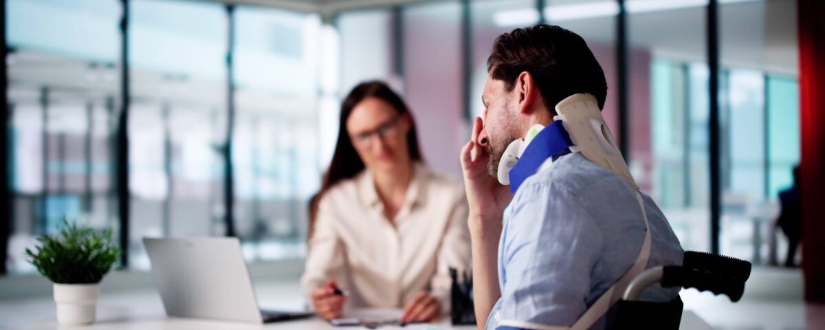 A man wears a neck brace and sits in a wheelchair next to a woman’s desk. He rubs his face with his hand.