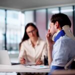 A man wears a neck brace and sits in a wheelchair next to a woman’s desk. He rubs his face with his hand.