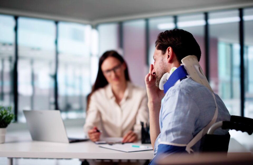 A man wears a neck brace and sits in a wheelchair next to a woman’s desk. He rubs his face with his hand.