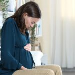 A pregnant woman in a teal shirt sits on a white couch. She frowns in pain as she holds on to her belly.