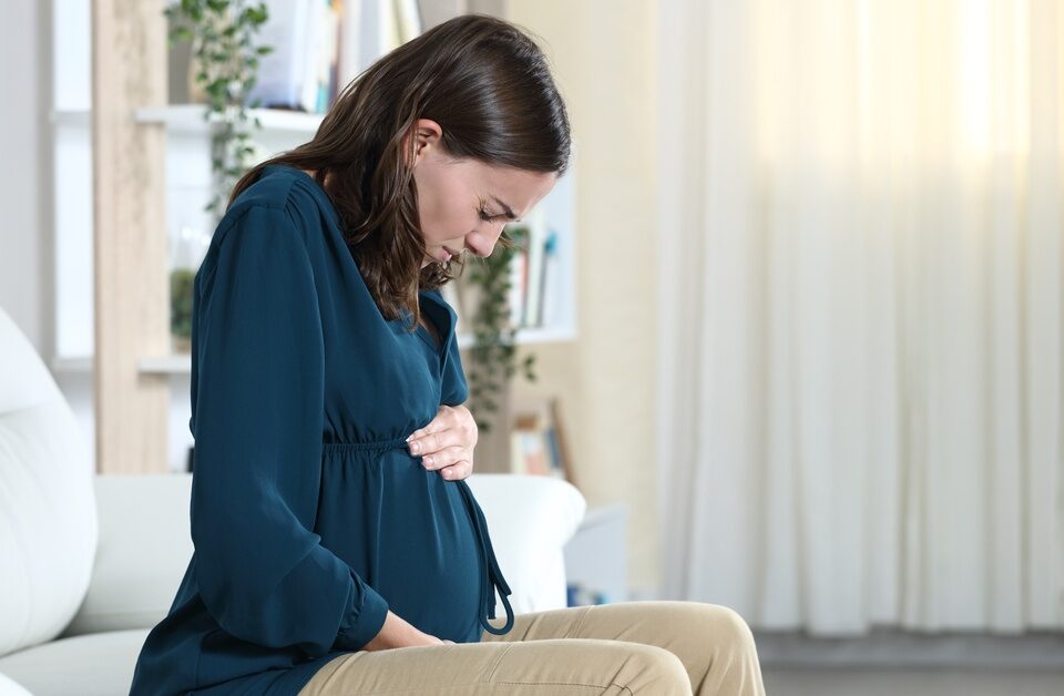 A pregnant woman in a teal shirt sits on a white couch. She frowns in pain as she holds on to her belly.