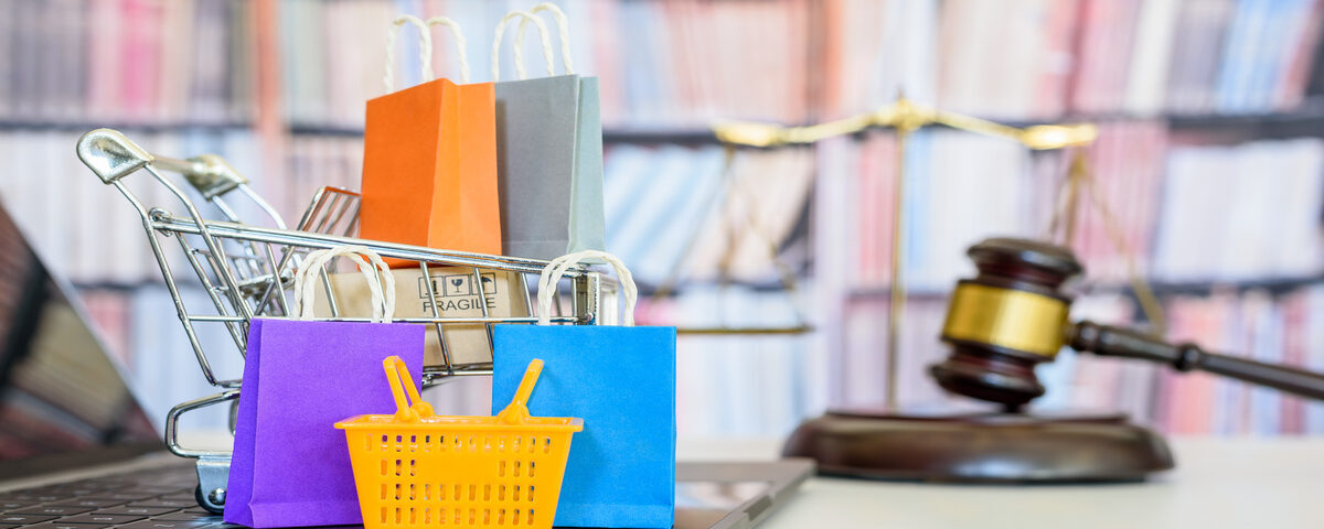 A miniature shopping cart filled with tiny bags and boxes on top of a laptop. There is a gavel and set of scales behind it.