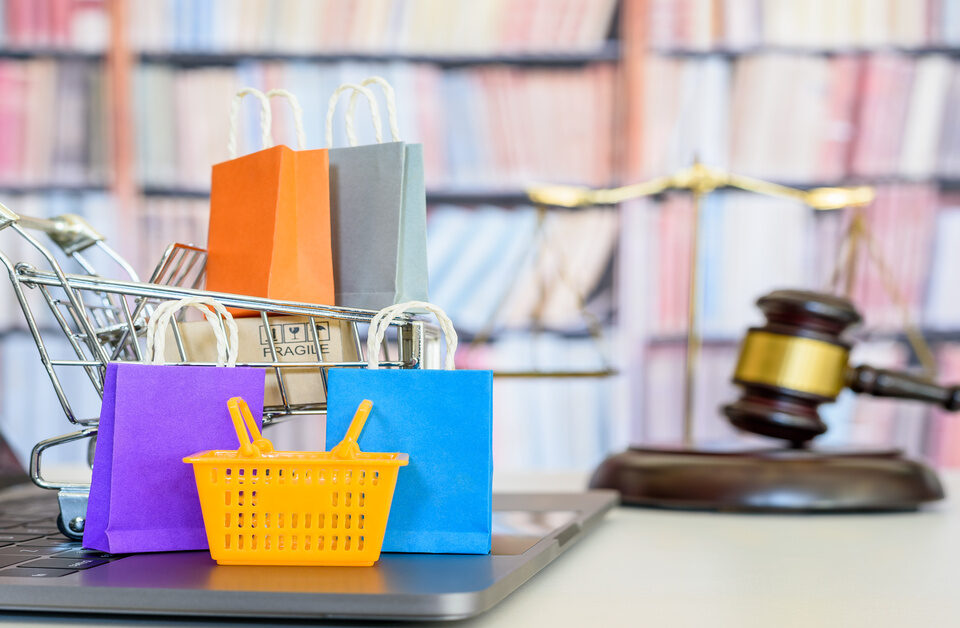 A miniature shopping cart filled with tiny bags and boxes on top of a laptop. There is a gavel and set of scales behind it.