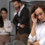 A young woman stands in an office and hides her face. In the background, three other people are looking at her.
