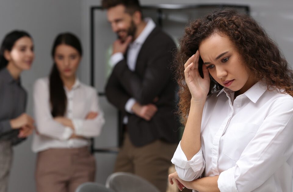 A young woman stands in an office and hides her face. In the background, three other people are looking at her.