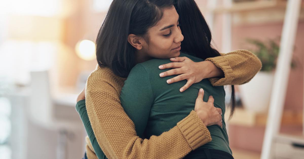 Two women hug each other. One woman is wearing a green blouse, and the other is wearing a brown sweater.