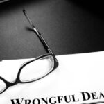 A close-up view shows scattered paper on a black surface with black glasses on top. The top paper reads "Wrongful Death."