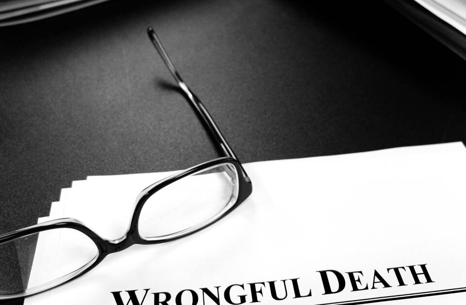 A close-up view shows scattered paper on a black surface with black glasses on top. The top paper reads "Wrongful Death."