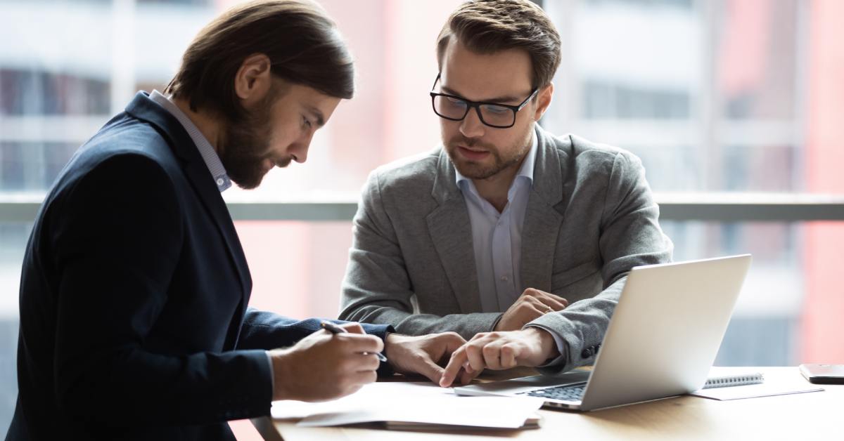 Two men dressed in professional business attire sit at a table looking at papers. An open laptop sits on the table.
