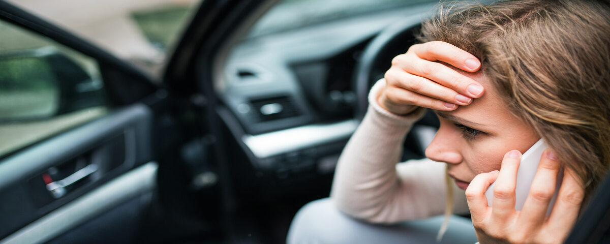 A stressed woman on a phone call, one hand pressed to her forehead, as she sits in the driver seat of her car with the door open.
