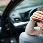 A stressed woman on a phone call, one hand pressed to her forehead, as she sits in the driver seat of her car with the door open.