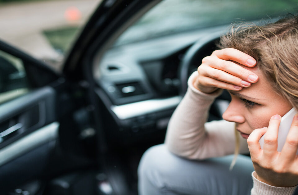 A stressed woman on a phone call, one hand pressed to her forehead, as she sits in the driver seat of her car with the door open.