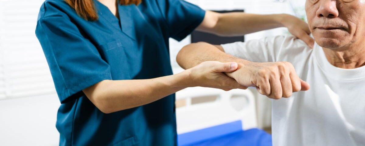 A female physiotherapist wearing blue scrubs is assisting an older adult male patient with rehabilitation exercises.