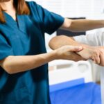 A female physiotherapist wearing blue scrubs is assisting an older adult male patient with rehabilitation exercises.