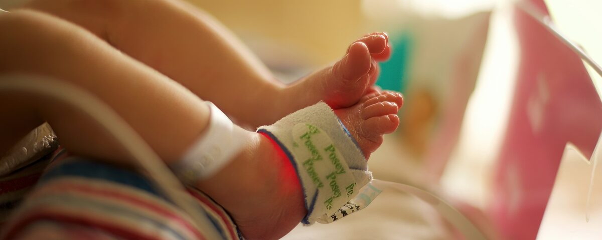 The feet of a newborn baby in a hospital setting. There is a pulse oximeter attached to one of the feet.