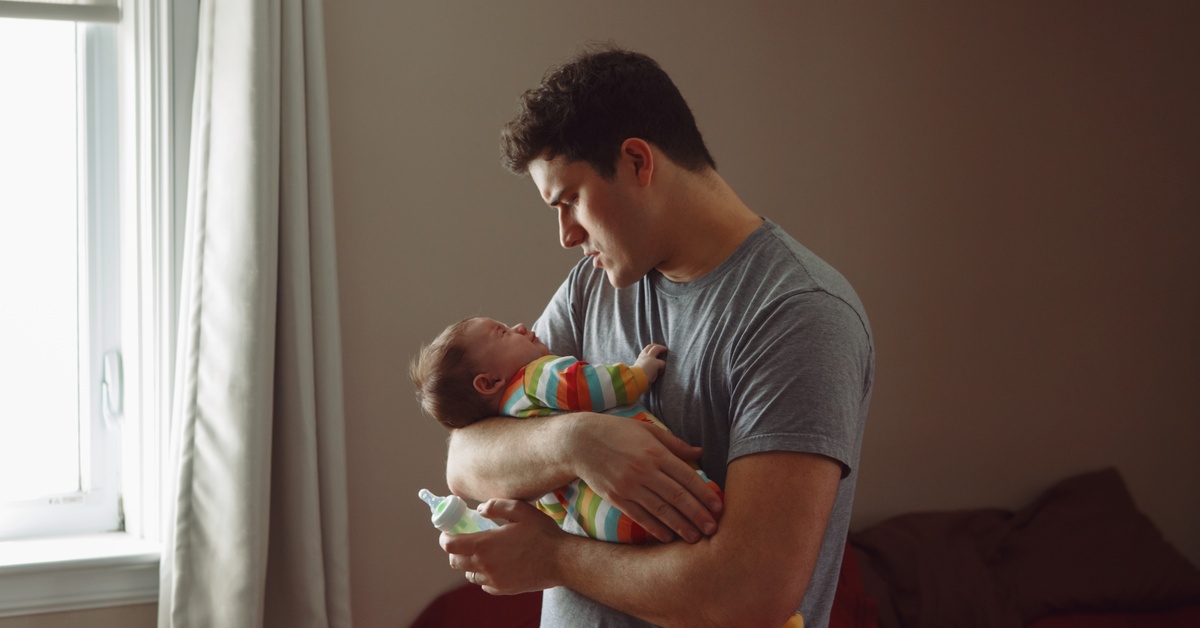 A man has a concerned look on his face while he holds a fussy newborn baby in a bedroom.